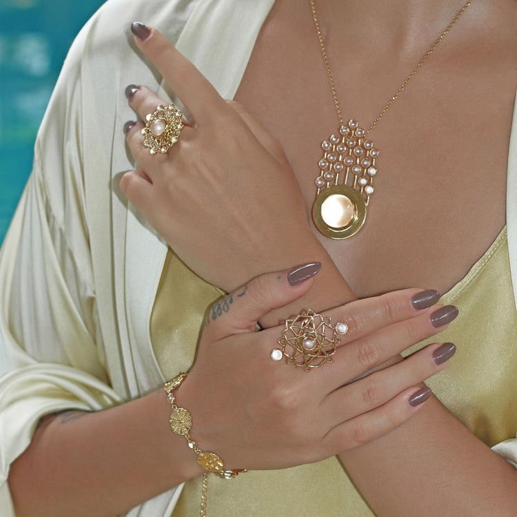 A woman poses with manicured nails, wearing Kaora Sandara Jewelry’s Ring Expansion gold plated statement ring, a bracelet, and a pendant necklace. She crosses her arms in a cream satin robe over a light gold dress.