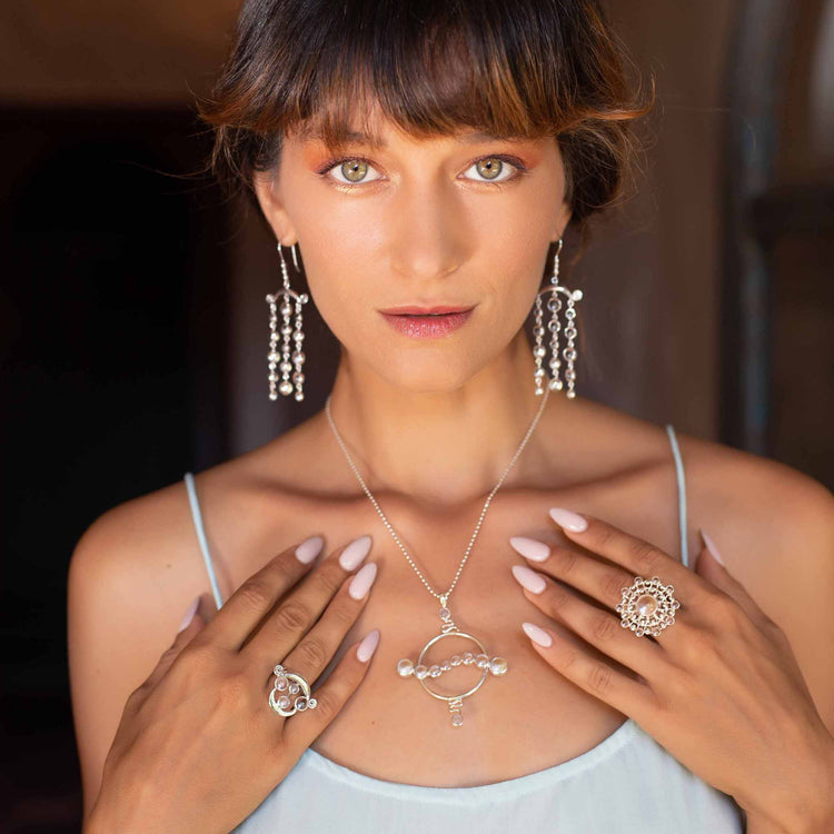 A woman with light brown hair and bangs wears the Kaora Sandara Jewelry Soul Potential Pendant—sterling silver with crystal and chalcedony—along with dangling pearl earrings, ornate rings, and a light blue spaghetti strap top as she gently touches her chest.