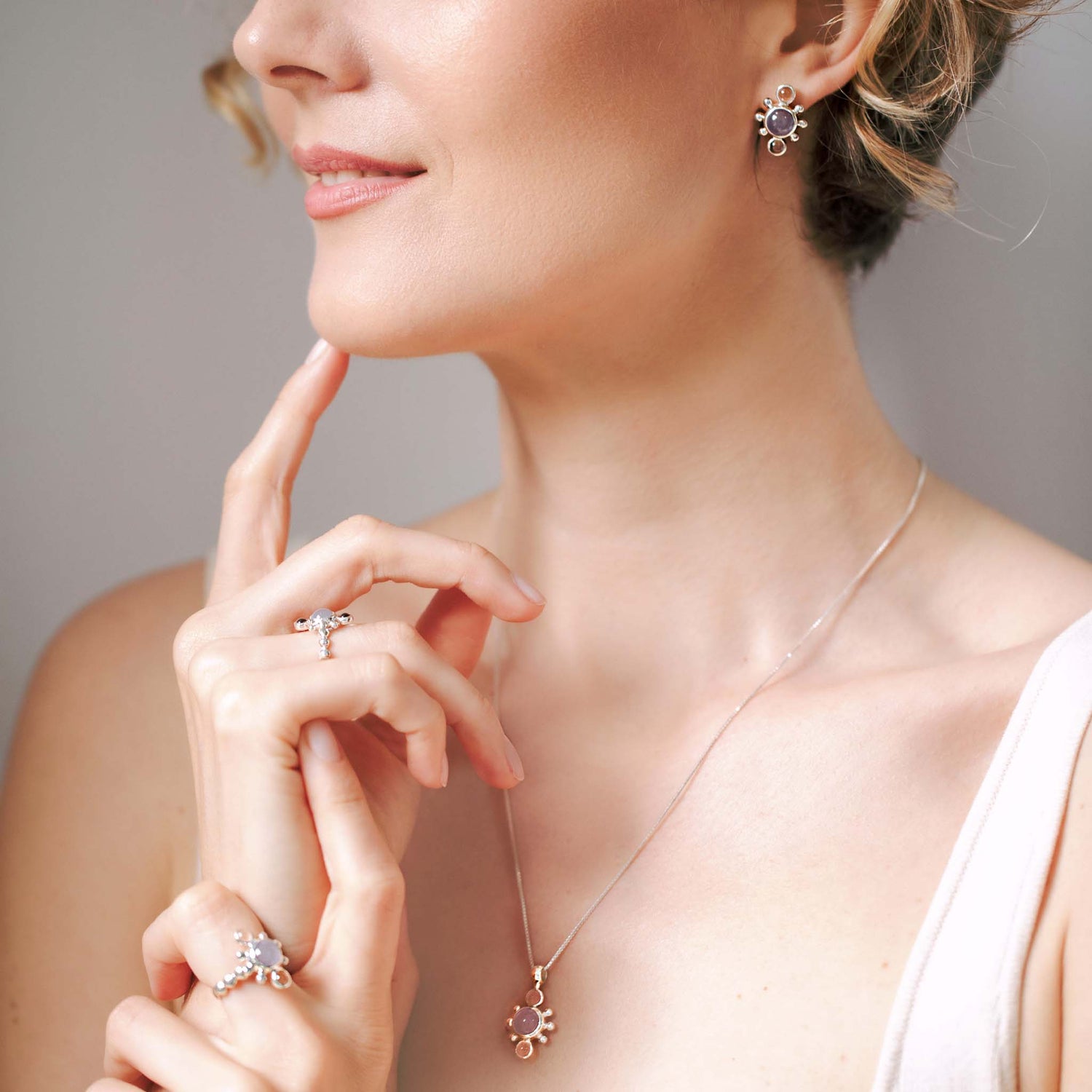 A woman with short, curly hair smiles softly, hand to chin, wearing Kaora Sandara Jewelry: Bliss Earhooks (Sterling Silver with Crystal & Chalcedony, Size L), a chalcedony pendant necklace, and Sterling Silver 925 rings with pearls and gemstones.