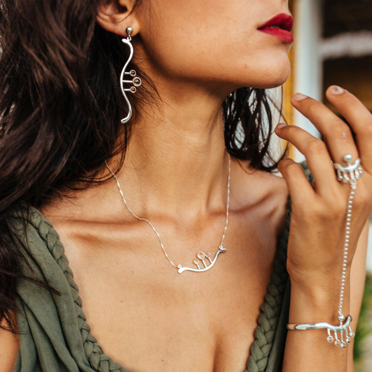 A woman wearing Kaora Sandara Jewelry's Silver Earstuds Beyond Fears with Crystal Spheres Gemstones, along with a matching necklace, bracelet, and chain-connected ring. She has red lipstick and a sleeveless deep green top.
