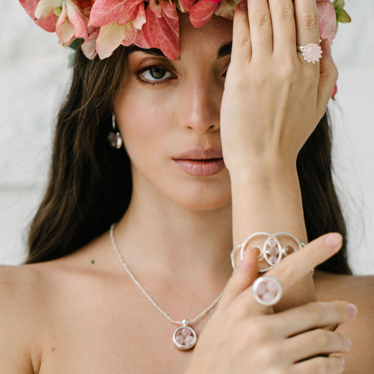 A woman with long dark hair and a pink flower crown wears Kaora Sandara Jewelry’s Silver Pendant Crystal Peace necklace with silver-rose quartz chain and rose quartz gemstones, covering one eye to display matching rings and bracelet as she gazes at the camera.