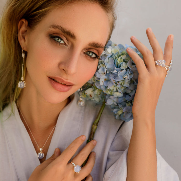 A woman with wavy blonde hair poses with a blue hydrangea, wearing the Kaora Sandara Jewelry Silver Pendant "I'M A Queen" with Chalcedony and a light, flowy top.