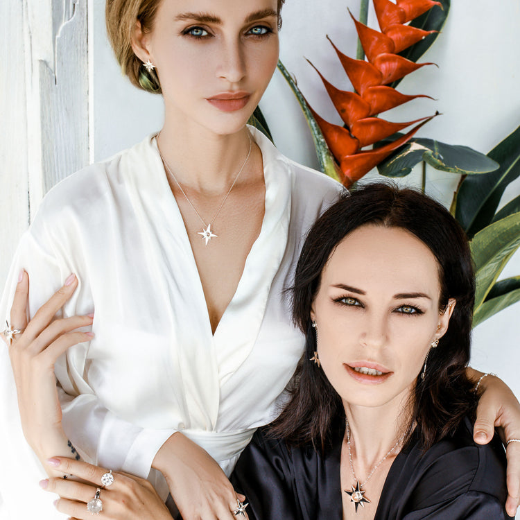 Two women, one with light hair and the other with dark hair, respectively dressed in white and black under a lush backdrop, are wearing Kaora Sandara Jewelry's Silver Pendant Magic Star with Crystal Sphere - Size XXS. The light-haired woman stands with her hand on the shoulder of the seated dark-haired woman as they both face the camera.