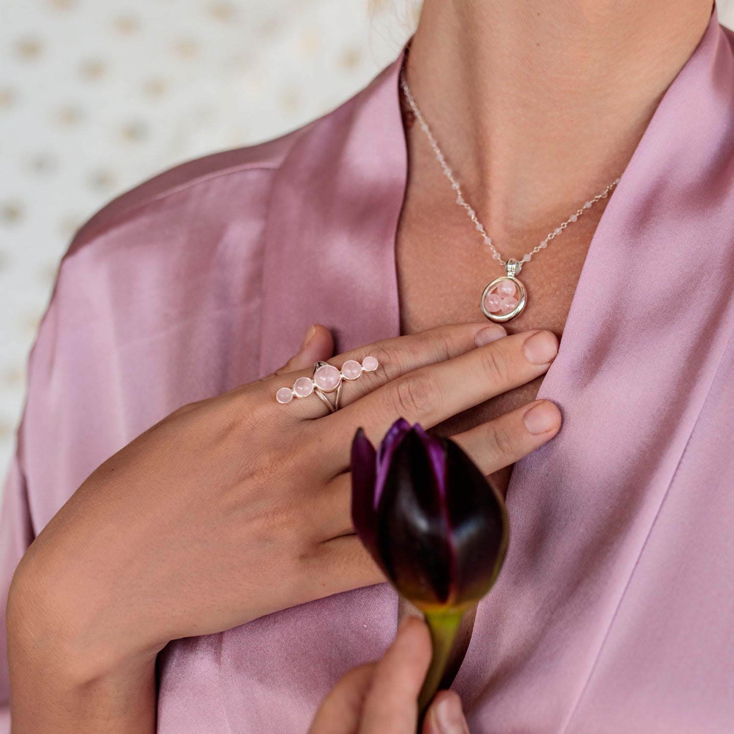 A woman in a pink satin robe holds a dark purple tulip and wears a silver necklace with a Rose Quartz pendant for universal love, plus Kaora Sandara Jewelry's Silver Ring Harmony with Rose Quartz Sphere Gemstones on her hand.