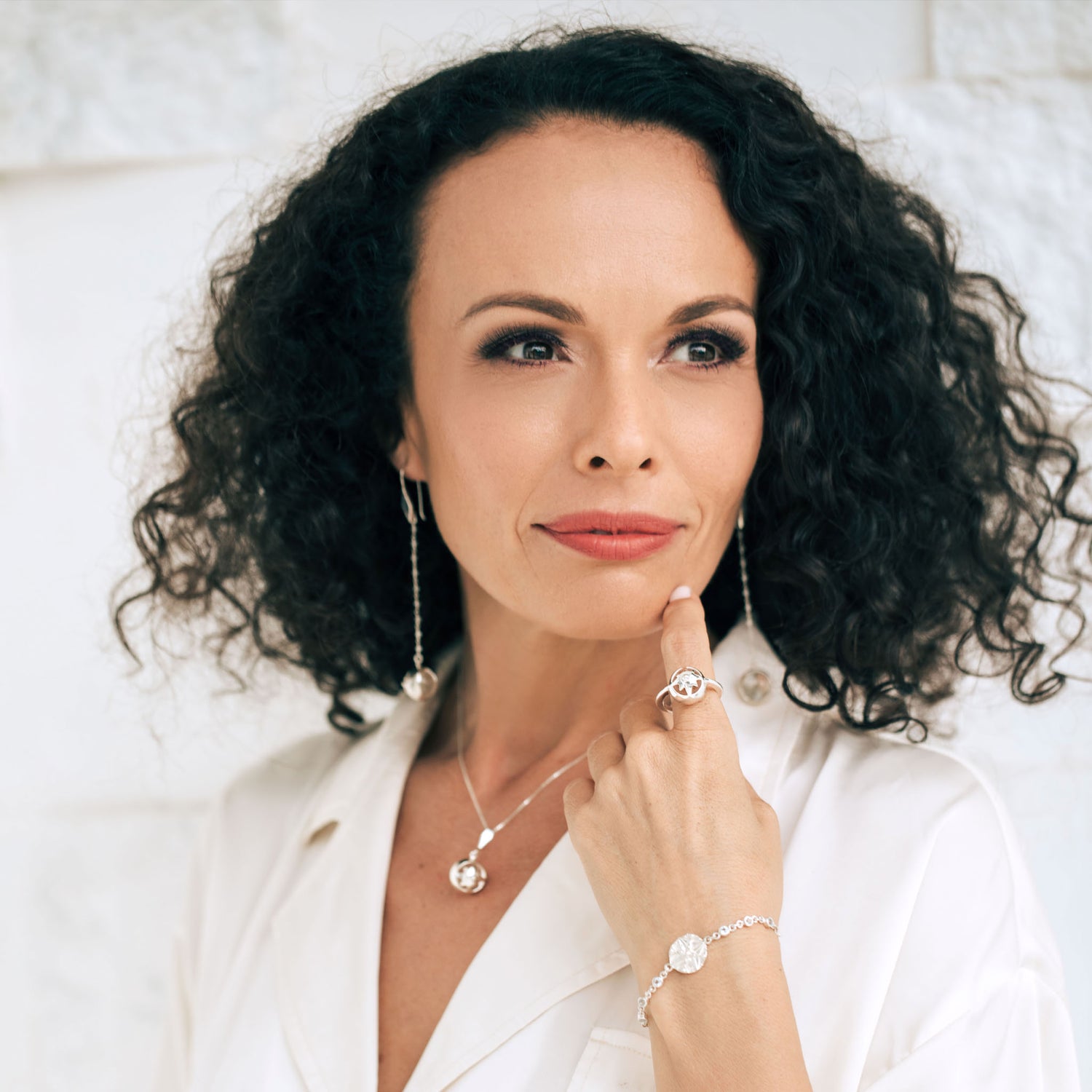 A woman with curly dark hair wears a white blouse and Kaora Sandara Jewelry, including the Magic Time Day Ring in sterling silver with a crystal gemstone, as she gazes off-camera and gently touches her chin with one finger.