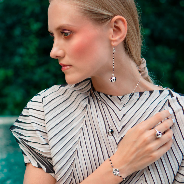 A woman with light hair, in a white and black striped top, poses before a blurred green backdrop wearing Kaora Sandara Jewelry’s Magic Time Night Ring—Sterling Silver with Black Agate Gemstone—alongside matching jewelry.