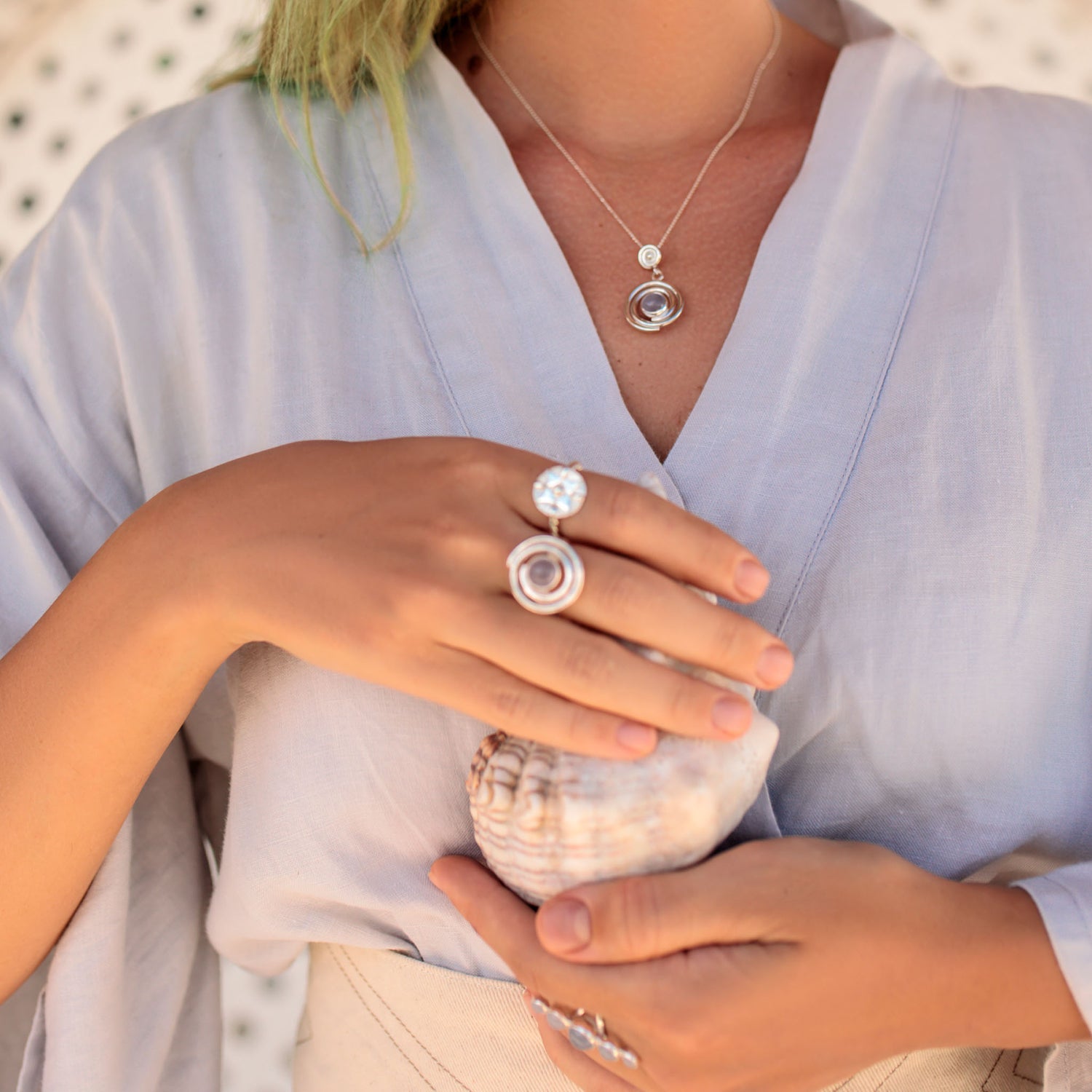 A person in a light blue robe with light green hair wears Kaora Sandara Jewelry's Silver Ring Open Spiral with Chalcedony (Size S) and holds a large seashell. The softly blurred background enhances the serene atmosphere.