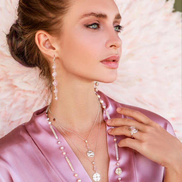 A woman in a silky pink robe wears Kaora Sandara Jewelry’s Sterling Silver 925 layered necklaces, dangling earrings, and the Silver Ring Open Spiral with Rose Quartz - Size S. She poses serenely against a feathery background, gazing to the side.