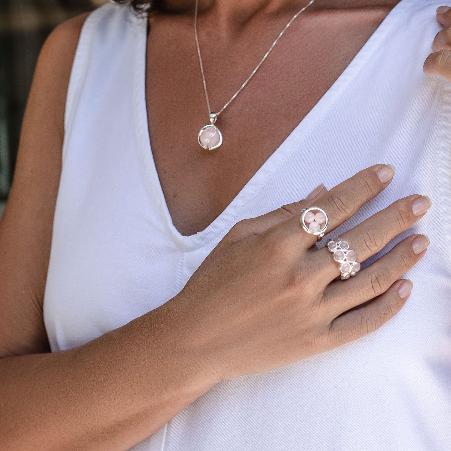 A woman in a white sleeveless top wears two Silver Ring The Dance with Rose Quartz and a matching pendant necklace by Kaora Sandara Jewelry, all crafted from sterling silver and adorned with light pink rose quartz stones. Her hand rests gently on her chest.