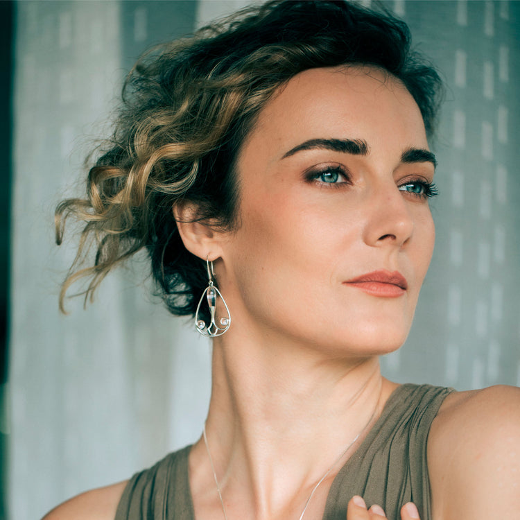 A woman with short, curly hair gazes thoughtfully, wearing a sleeveless taupe top and Kaora Sandara Jewelry’s Silver Earhooks Self-Worth – Crystal Gemstone. She stands before a softly lit sheer curtain backdrop.