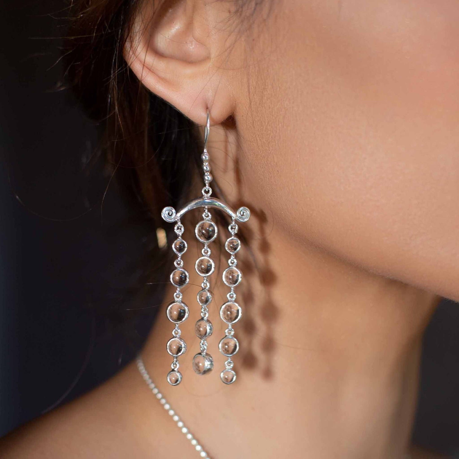 Close-up of a person wearing Kaora Sandara Jewelry's Magic Path Earhooks—Sterling Silver earrings with crystal gemstones. Three vertical rows of circular links hang from a curved bar; smooth skin and part of a necklace are visible.