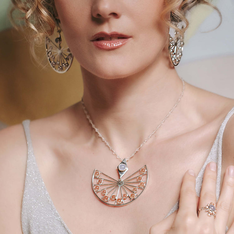 A woman in a sparkling silver dress models Kaora Sandara Jewelry’s Higher Self Pendant—Sterling Silver with Crystal & Chalcedony—paired with matching ornate earrings and a decorative ring. Her lips are glossy, hair styled in soft curls.