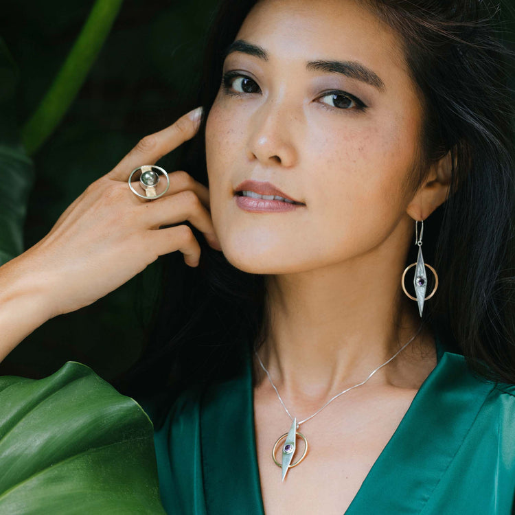 A woman in a green top poses with her hand near her face, wearing Kaora Sandara Jewelry’s Intuition Earhooks—sterling silver and gold-plated earrings with amethyst. Circular and pointed shapes stand out against a leafy background.