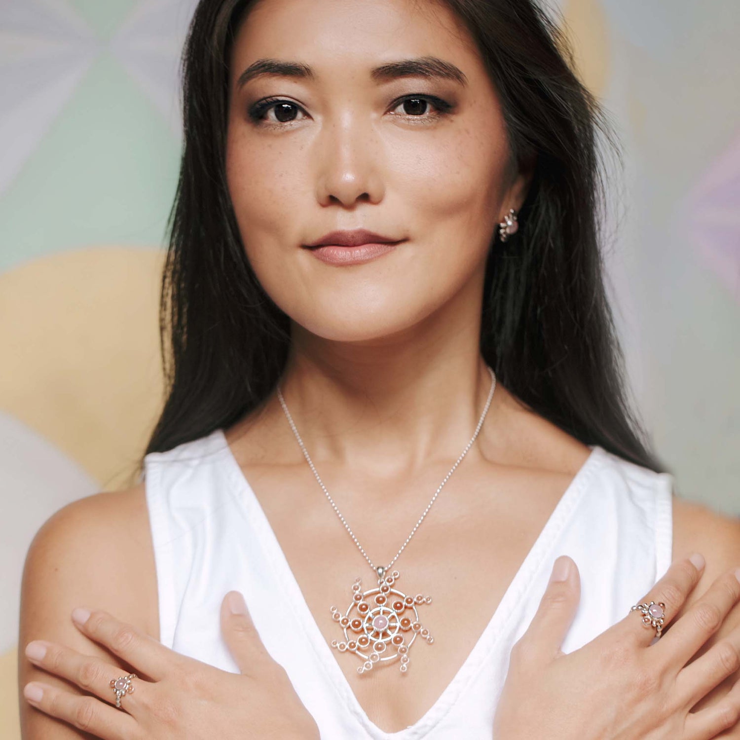 A woman with long dark hair wears a white sleeveless top and Kaora Sandara Jewelry's Perfection of Life Pendant—Sterling Silver with Crystal & Rose Quartz—plus matching earrings and rings, standing with hands crossed and smiling gently.