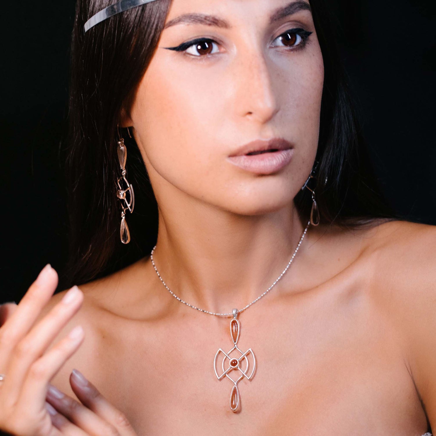 A woman with long dark hair wears the Kaora Sandara Jewelry Soul Guidance Pendant—Sterling Silver with Crystal—and matching earrings. She has natural makeup, winged eyeliner, and gazes off-camera against a dark background.