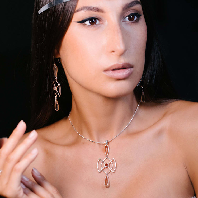 A woman with long dark hair wears the Kaora Sandara Jewelry Soul Guidance Pendant—Sterling Silver with Crystal—and matching earrings. She has natural makeup, winged eyeliner, and gazes off-camera against a dark background.