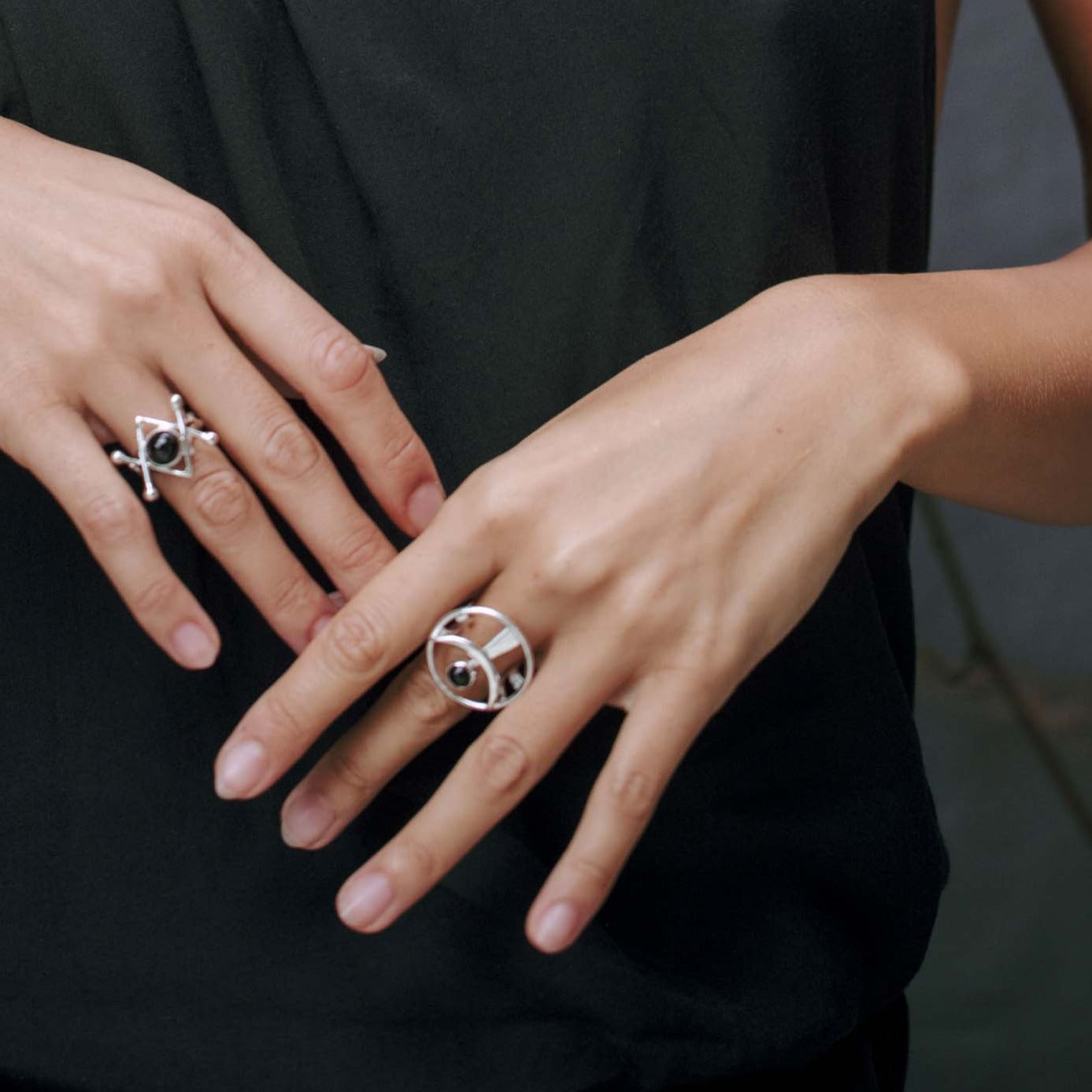 A person wearing a black sleeveless top displays their hands adorned with two Kaora Sandara Jewelry "Releasing From the Deepest Fears" rings in Sterling Silver with Black Agate (Size L). The background is neutral and blurred.