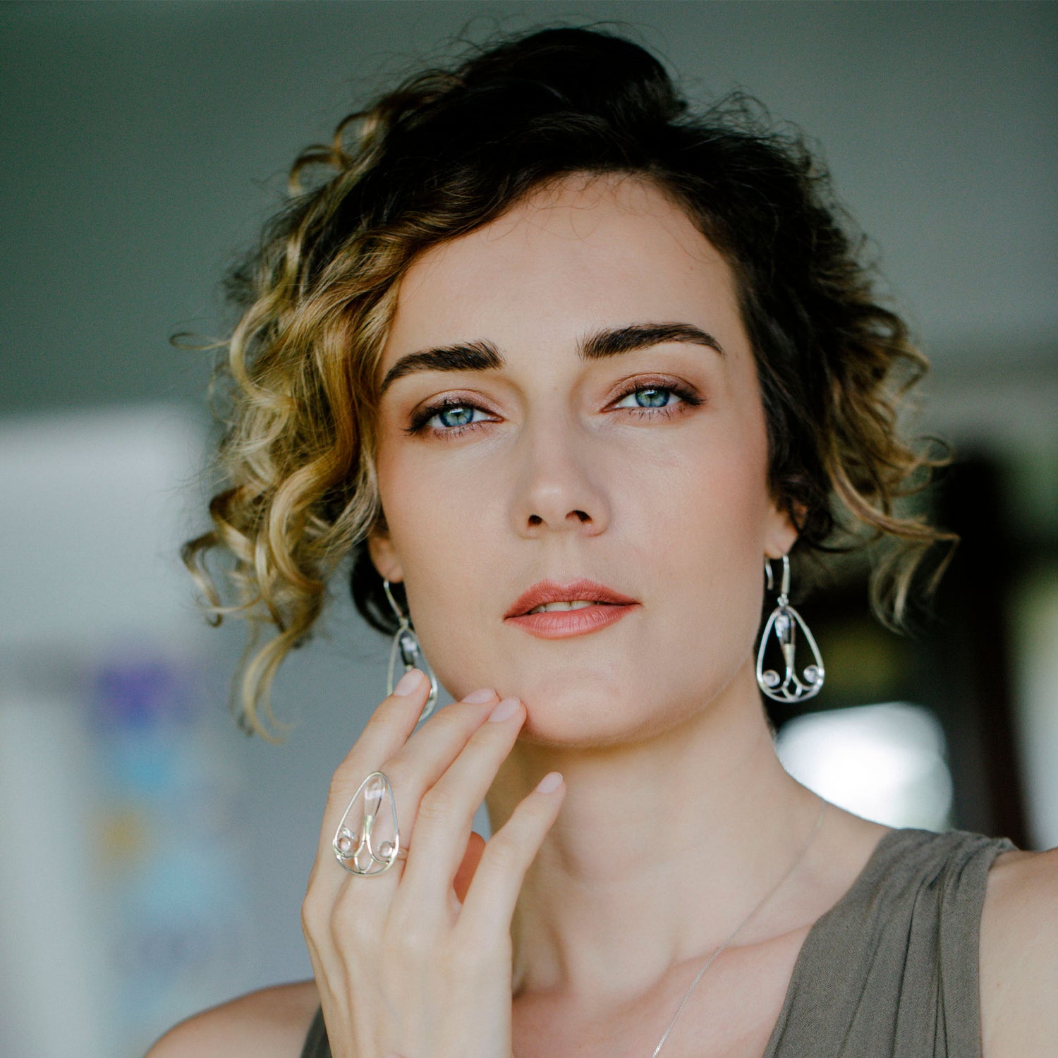 A woman with short, curly hair and blue eyes wears Kaora Sandara Jewelry’s Self-Worth Ring—Sterling Silver with Crystal. She gently touches her chin, gazing calmly at the camera as a softly blurred background highlights her elegance.