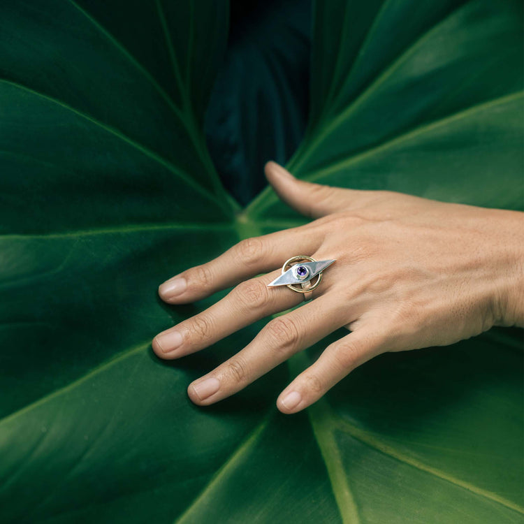 A hand adorned with the Kaora Sandara Jewelry Intuition Ring—Sterling Silver & Gold-Plated with Amethyst—rests on a large green tropical leaf, fingers gently splayed across its surface.