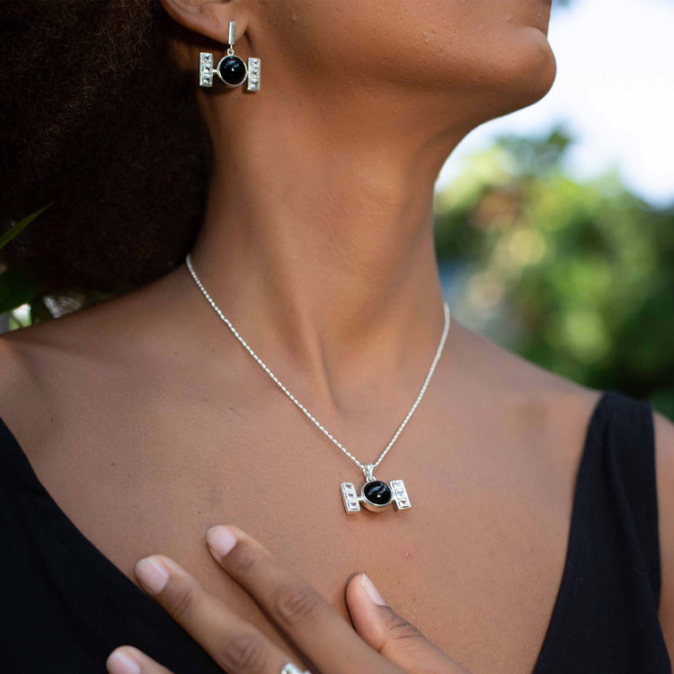A woman outdoors in a sleeveless black top wears Kaora Sandara Jewelry’s Health Formula Pendant—Sterling Silver with Black Agate & Crystal—paired with matching TIE fighter-shaped earrings and a silver ring, greenery blurred in the background.
