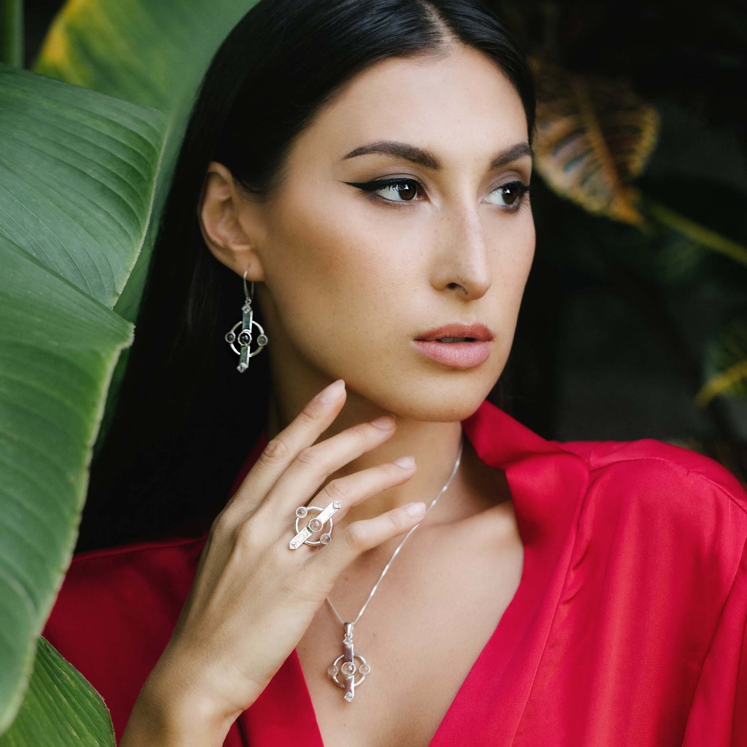 A woman in a red blouse poses among green leaves, wearing Kaora Sandara Jewelry's Inner Clarity Earhooks—Sterling Silver with Crystal Gemstones. With long dark hair, she gazes thoughtfully to the side, embodying clarity.