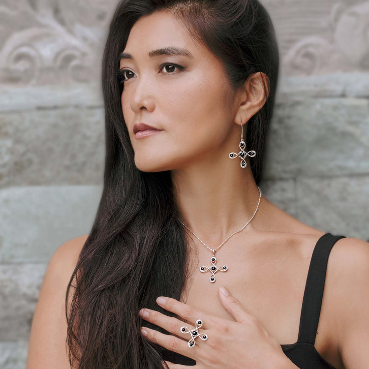 A woman with long dark hair wears a black dress and Kaora Sandara Jewelry's Programs Clearing Earhooks—sterling silver earrings featuring black agate. She gazes thoughtfully to the side, hand resting gently on her chest.