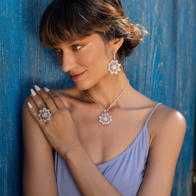 A woman in a lavender dress poses against a blue wooden wall, wearing Kaora Sandara Jewelry's Source of Love Pendant in sterling silver with crystal and rose quartz. She gazes to the side, her hand resting on her shoulder.