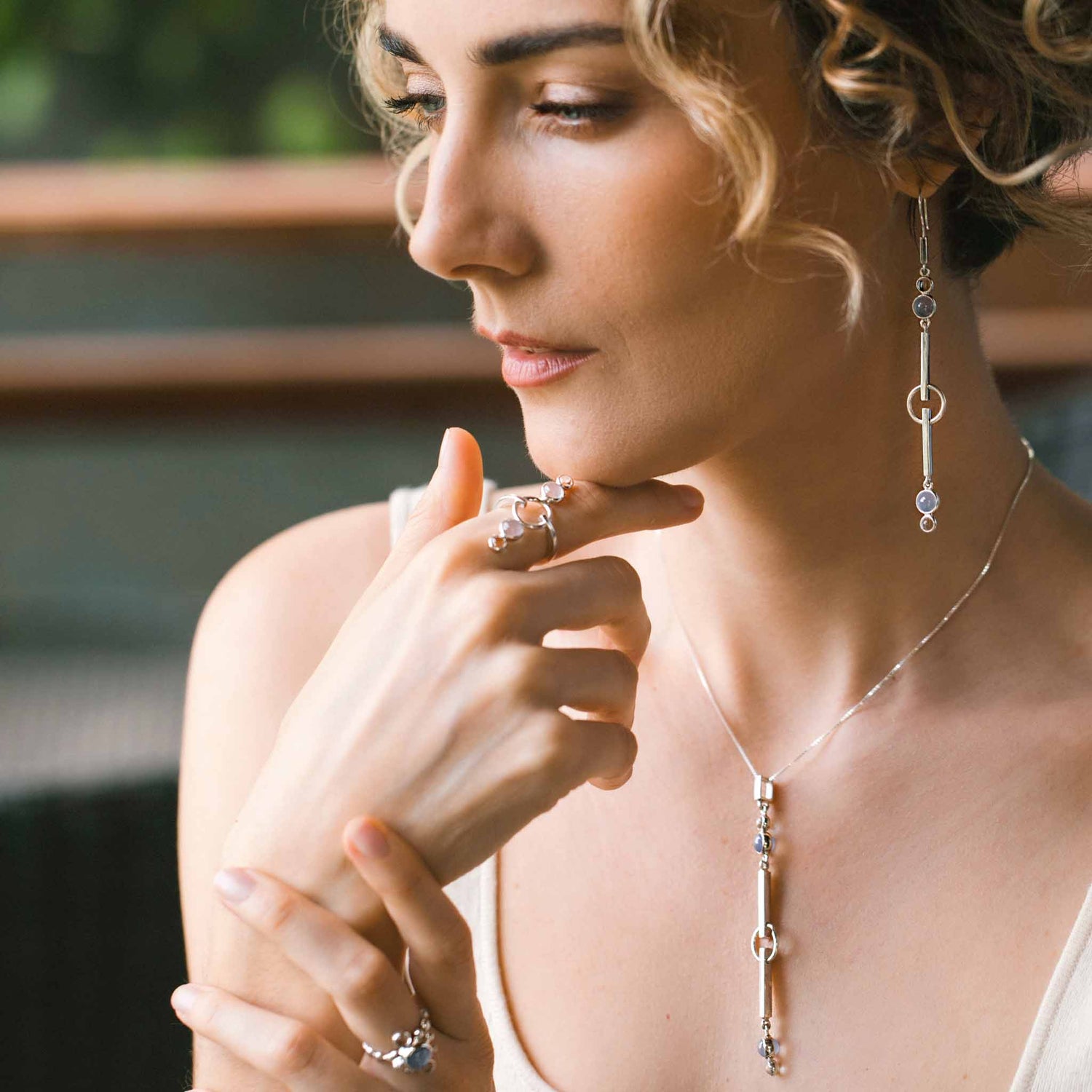 A woman with curly hair gazes thoughtfully, radiating tranquility as she wears Kaora Sandara Jewelry’s Silver Earhooks State—elegant earrings adorned with crystal and chalcedony gemstones in delicate circular settings.