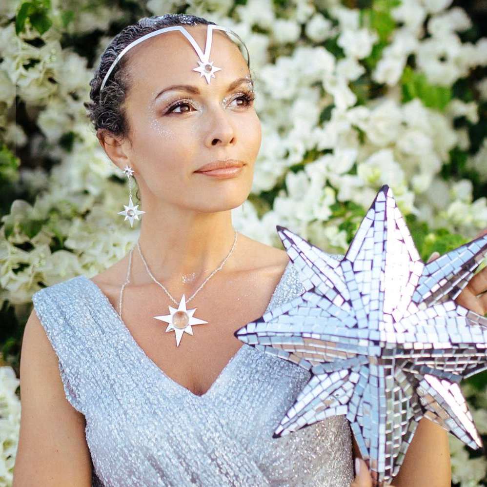 A woman wearing Kaora Sandara Jewelry's Silver Earstuds on Chain Magic Star with Crystal Sphere Gemstones sparkles in a shimmering silver dress, mirrored star sculpture in hand, as white flowers bloom behind her ethereal elegance.