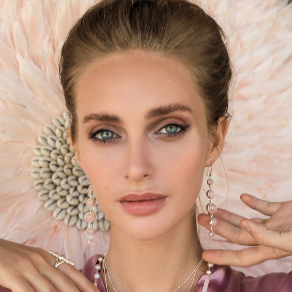 A woman with light eyes and styled brown hair models Kaora Sandara Jewelry’s Harmony Earhooks—sterling silver with rose quartz. She wears delicate rings, layered necklaces, a soft pink top, and natural, polished makeup by a large pale flower.