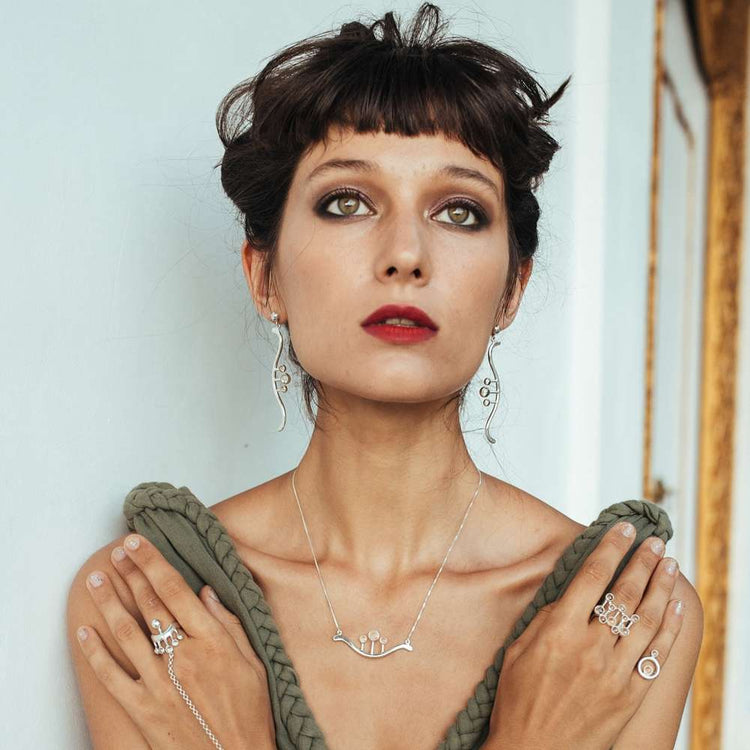 A woman with short dark hair and bangs wears the Kaora Sandara Jewelry Silver Pendant Beyond Fears on a thin 45cm square chain with crystal, paired with dramatic eye makeup, red lipstick, and an off-shoulder green top.