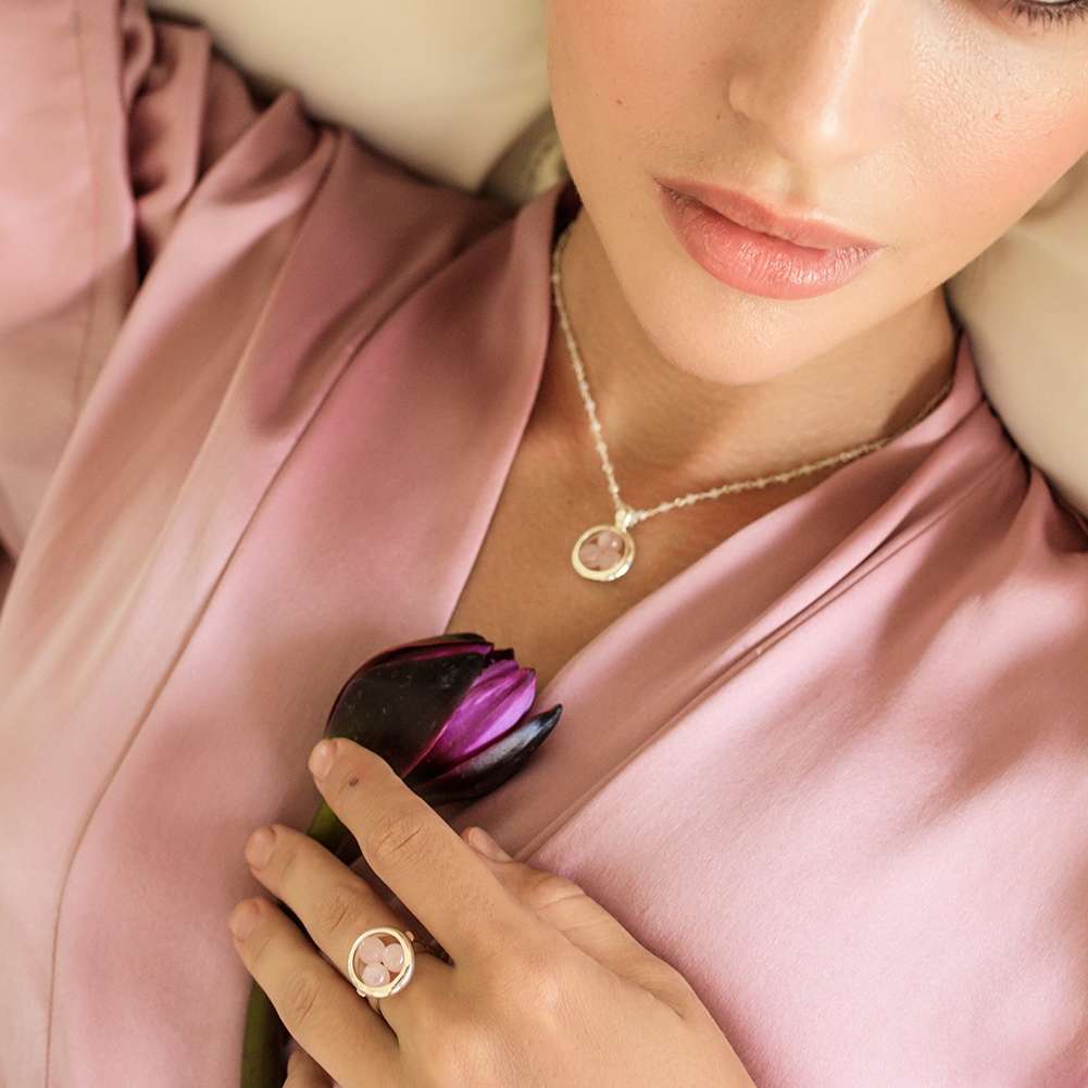 A woman in a pink satin robe holds a dark purple tulip and wears the Kaora Sandara Jewelry Silver Ring Crystal Peace with Rose Quartz, size S, lying down with soft makeup and natural lips.