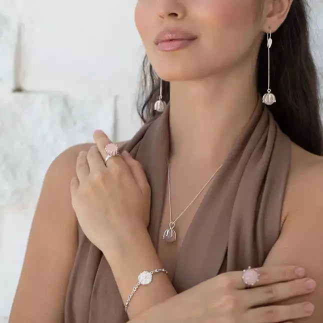 A woman in a sleeveless brown top models Kaora Sandara Jewelry’s Silver Pendant I'M A Queen Rose Quartz, along with matching rings, a bracelet, and long drop earrings. She poses with crossed arms against a light background.